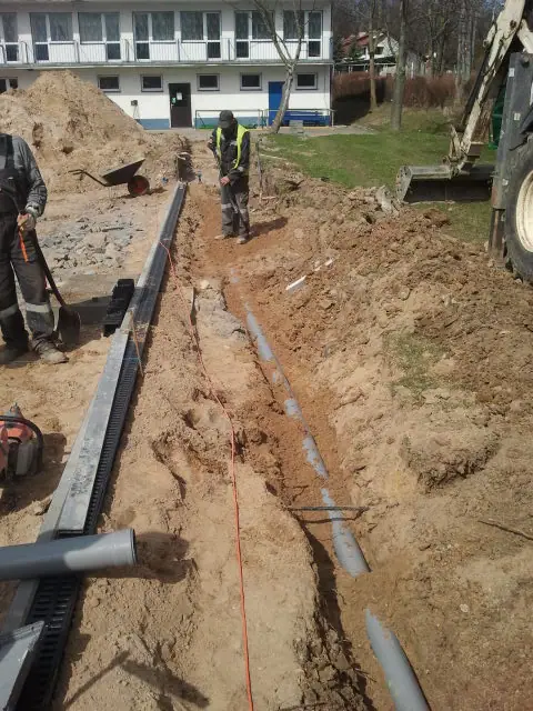 Pracownicy przy koparce montują odwodnienie liniowe wzdłuż wykopu przed budynkiem ośrodka