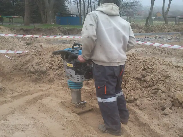 Robotnik zagęszcza podłoże ubijakiem spalinowym za taśmą ostrzegawczą na terenie boiska