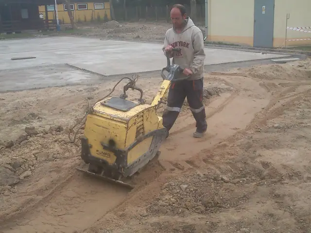 Pracownik prowadzi zagęszczarkę płytową po świeżo wylanej betonowej płycie boiska