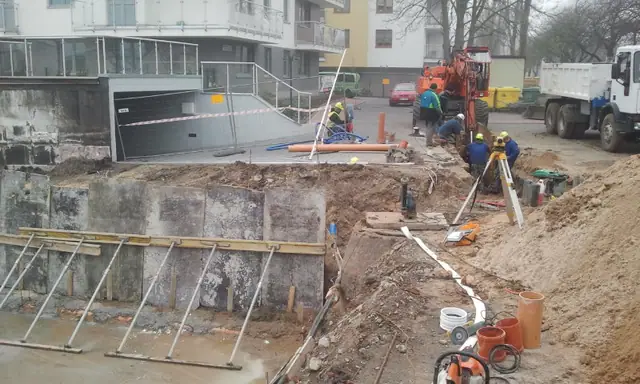 Geodeta z niwelatorem mierzy wykop obok ułożonych rur w pobliżu istniejącego budynku