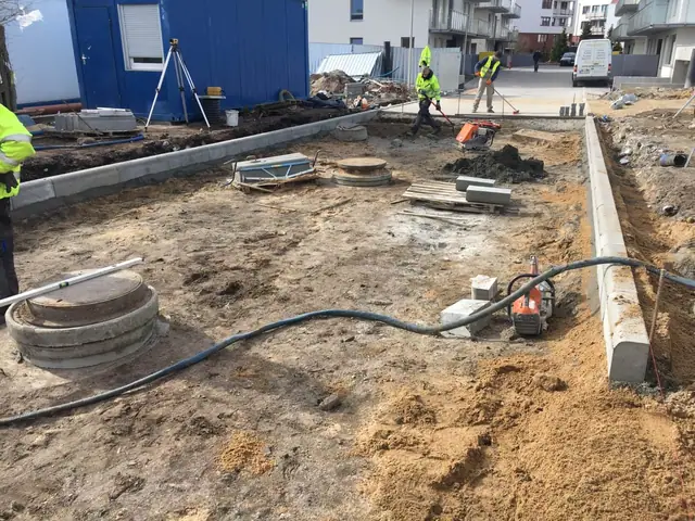 Robotnicy układają betonowy krawężnik przy studzience kanalizacyjnej obok kontenera socjalnego