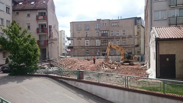 Koparka rozbija pozostałości ściany, obok rośnie hałda gruzu z rozbiórki