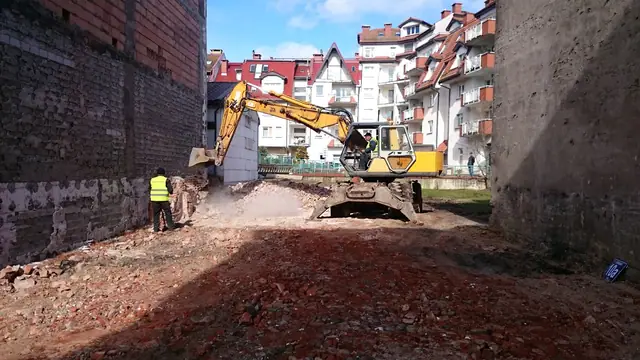 Koparka rozbiera fragment ściany kamienicy w otoczeniu miejskiej zabudowy