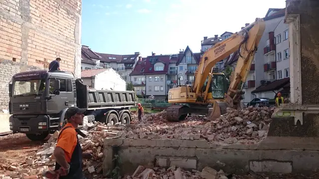 Koparka ładuje gruz z rozbiórki na samochód ciężarowy przy resztkach murów
