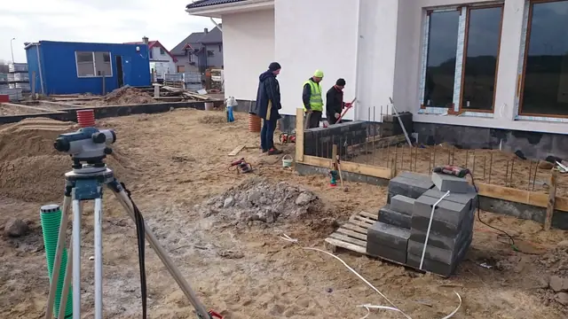 Geodeta z niwelatorem mierzy teren przy murowanym fundamencie ogrodzenia posesji