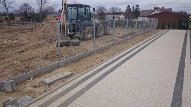 Koparko-ładowarka pracuje przy ogrodzeniu z siatki na tyłach posesji jednorodzinnej