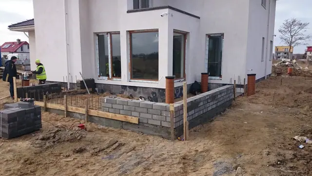 Murowanie niskiego ogrodzenia z pustaków wokół narożnika domu, widoczne rury drenażowe