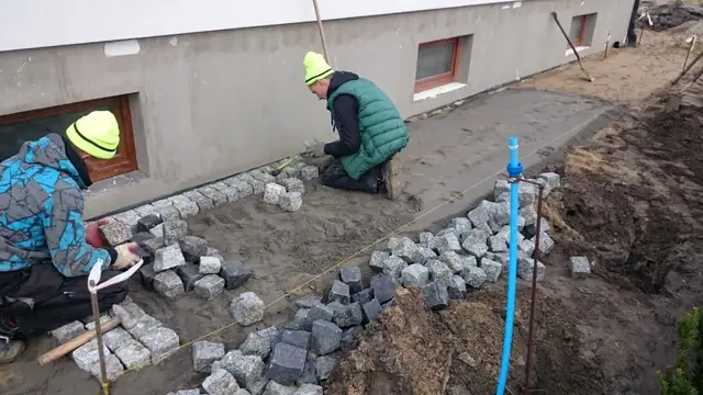 Ekipa brukarska układa kostkę granitową wzdłuż elewacji budowanego domu jednorodzinnego
