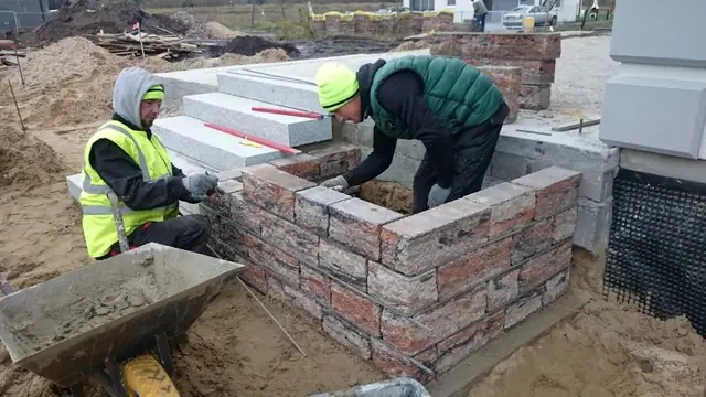 Murarze wznoszą narożny murek z cegły klinkierowej przy schodach tarasu