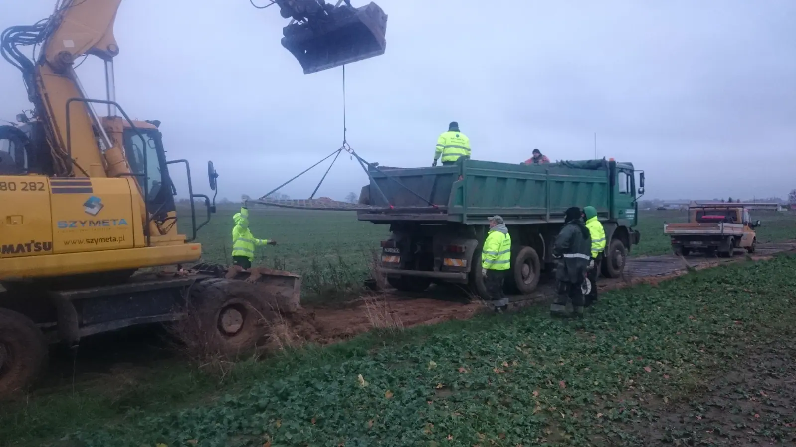 Koparka Komatsu firmy Szymeta rozładowuje kruszywo z wywrotki na rozjeżdżonej drodze polnej