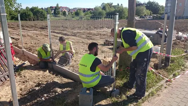 Dwóch pracowników montuje siatkę na stalowym słupku ogrodzeniowym przy chodniku z kostki