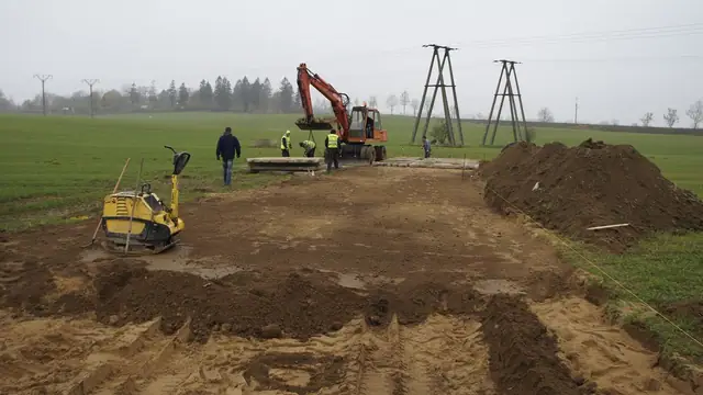 Pracownicy i koparka układają betonowe płyty drogi technicznej wśród pól