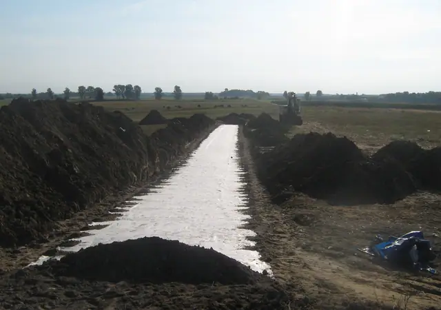 Pas białej geowłókniny rozłożony w wykopie otoczonym czarną ziemią, w tle sprzęt budowlany