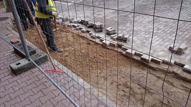 Robotnik grabi podsypkę wzdłuż nowo ułożonego krawężnika za siatką ogrodzeniową