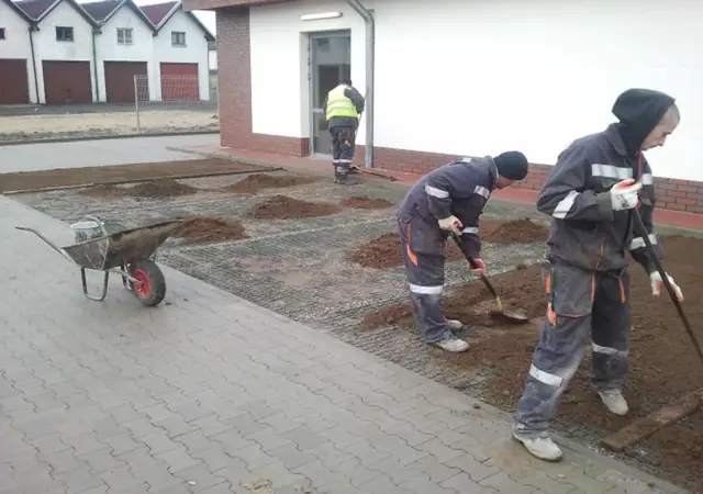 Dwóch robotników grabi ziemię pod trawnik przy wejściu do budynku, obok taczka