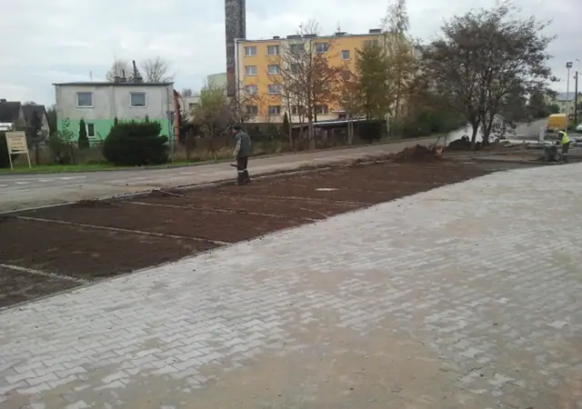 Robotnik wyrównuje ziemię pod trawnik na dużym placu, w tle bloki mieszkalne