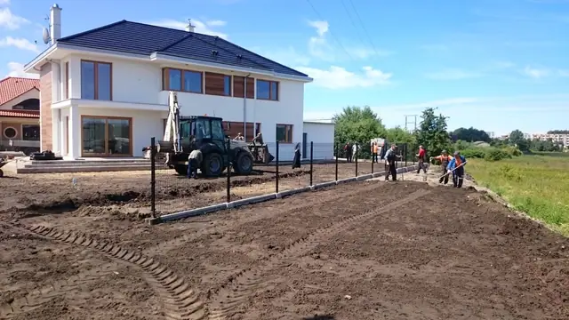 Koparko-ładowarka i robotnicy wytyczają teren pod trawnik ogrodzony płotkiem przy nowym domu
