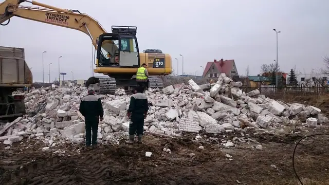 Pracownicy nadzorują załadunek gruzu z rozbiórki, obok koparka Komatsu PC210 i wywrotka