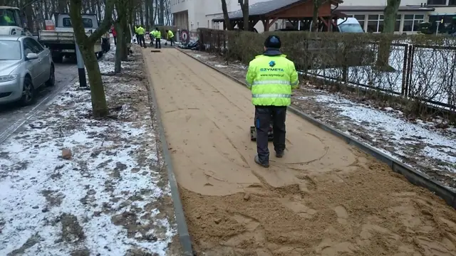 Pracownik w kurtce Szymeta rozprowadza podsypkę piaskową na trasie przyszłego chodnika w śniegu
