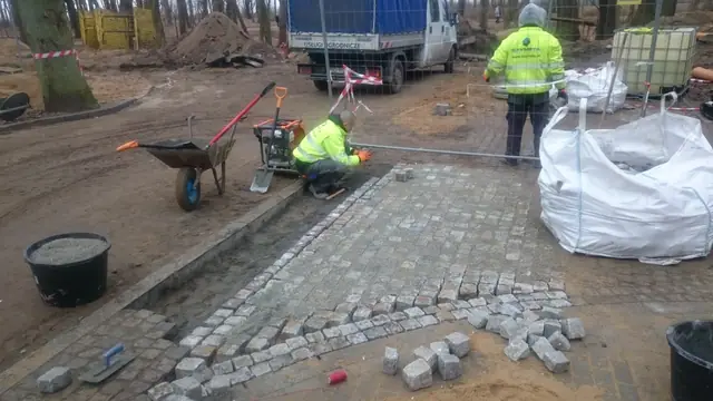 Pracownik układa krawężnik z kostki granitowej obok taczki i zagęszczarki płytowej