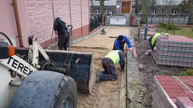 Robotnicy układają czerwoną kostkę brukową na obrzeżu trawnika obok koparki