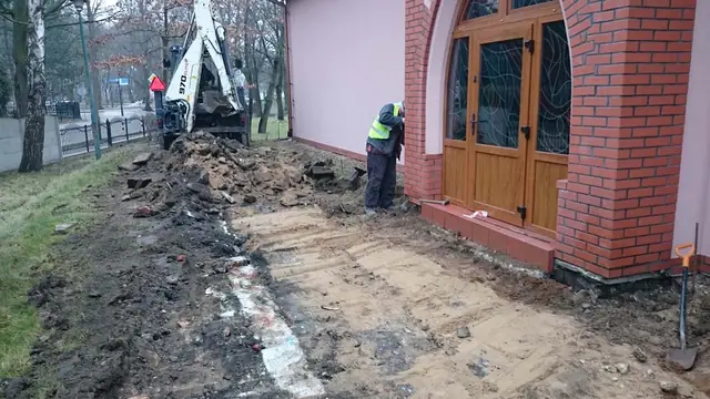 Mini koparka i robotnik pracują przy wykopie tuż obok głównych drzwi kościoła
