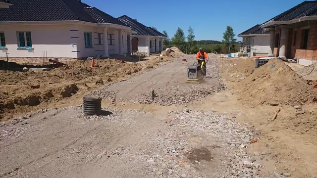 Pracownik zagęszcza żwirową drogę zagęszczarką obok odcinka rury drenażowej przy domach