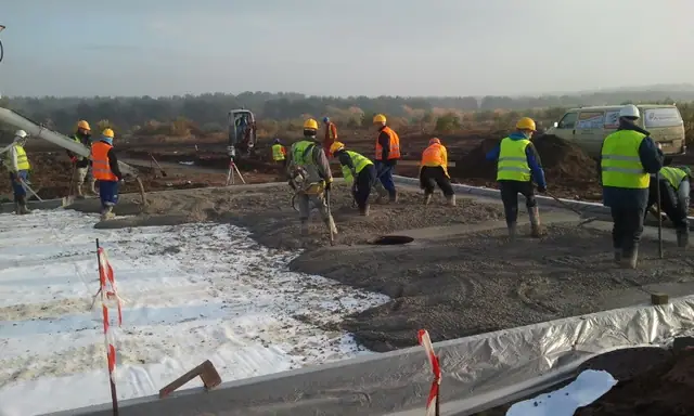 Liczna ekipa wykańcza rozległą płytę betonową obramowaną izolacją i taśmą ostrzegawczą
