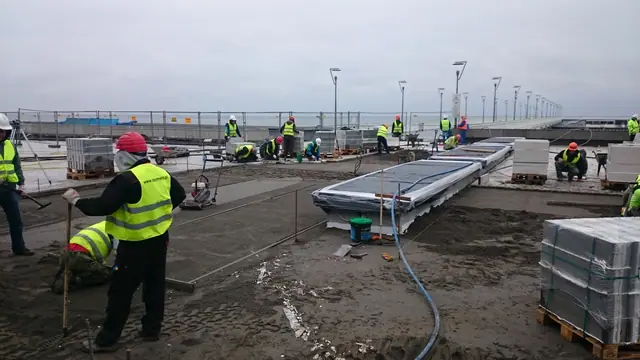 Grupa robotników układa długie elementy prefabrykowane na promenadzie nadmorskiej przy molo w Kołobrzegu