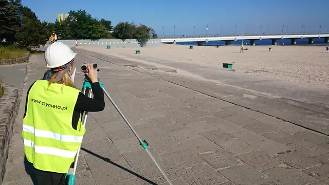 Pracownik w kasku z tyczką geodezyjną wykonuje pomiary na plaży przed remontem terenu przy molo w Kołobrzegu