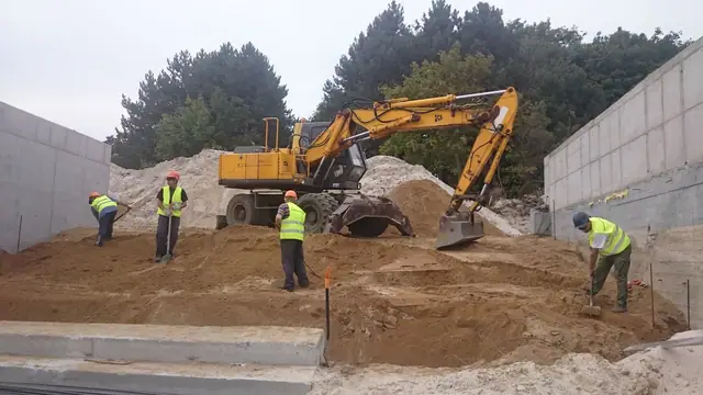 Koparka i robotnicy pracują przy murze oporowym obok wykopu z widoczną instalacją przy molo w Kołobrzegu