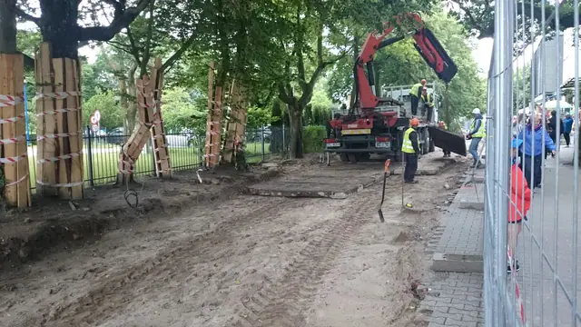 Koparko-ładowarka pracuje za ogrodzeniem budowy przy wbijaniu drewnianych pali podczas prac ziemnych przy molo
