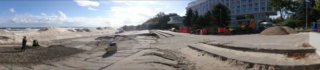 Koparka formuje wał piaszczysty na tle hotelu i betonowych schodów w trakcie budowy promenady w Kołobrzegu