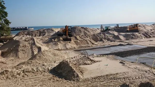 Koparka pracuje w wykopie fundamentowym otoczonym kopcami piasku przy budowie mola w Kołobrzegu