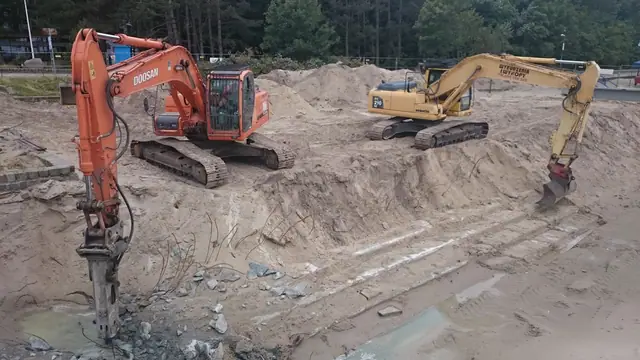 Dwie koparki gąsienicowe z młotami hydraulicznymi kruszą stary fundament wśród piasku przy molo w Kołobrzegu