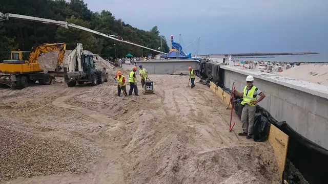 Koparko-ładowarki układają materiał wzdłuż nowego muru oporowego zabezpieczonego workami z piaskiem przy molo