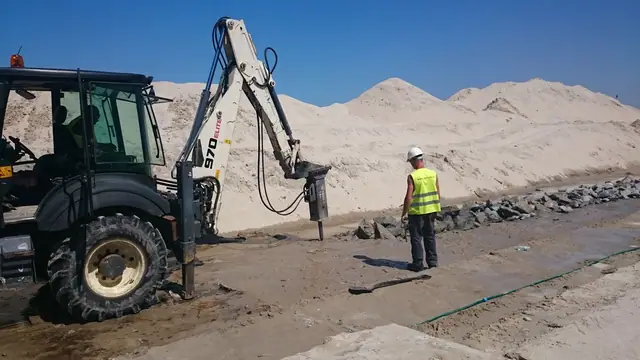 Koparko-ładowarka z młotem hydraulicznym kruszy stary fundament w piasku podczas rozbiórki przy molo