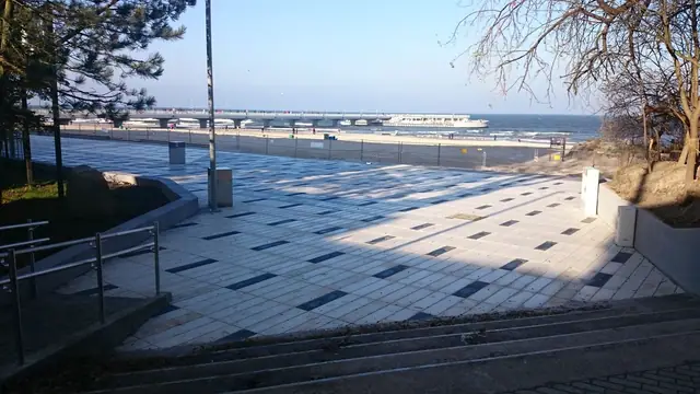 Narożnik placu przy molo w Kołobrzegu z rabatą żwirową, niewielkimi krzewami i krawędzią drewnianej ławki