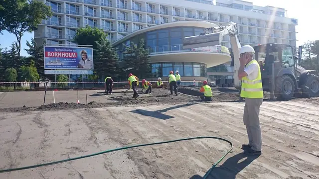 Pracownik z niwelatorem wykonuje pomiary geodezyjne na tle hotelu podczas budowy placu przy molo w Kołobrzegu
