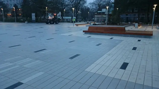Szeroki plac przy molo w Kołobrzegu oświetlony zapalającymi się latarniami, w tle widoczny ciągnik