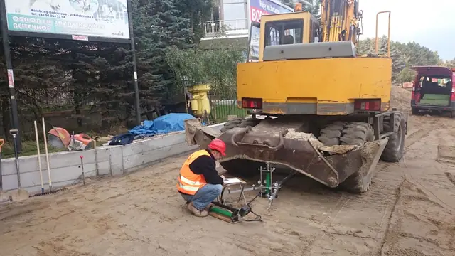 Pracownik klęczący przy koparce wykonuje przegląd sprzętu obok nowego muru oporowego przy molo w Kołobrzegu