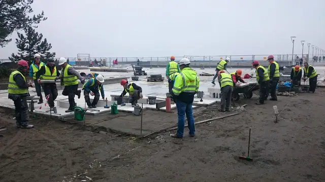Grupa robotników pracuje na placu przy budynku z widokiem na morze i molo w Kołobrzegu