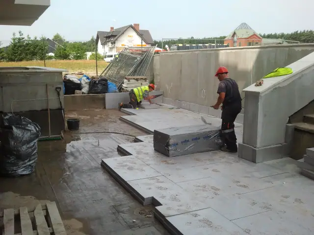 Robotnicy montują prefabrykowane elementy betonowe na wybrukowanej podstawie przy murku oporowym