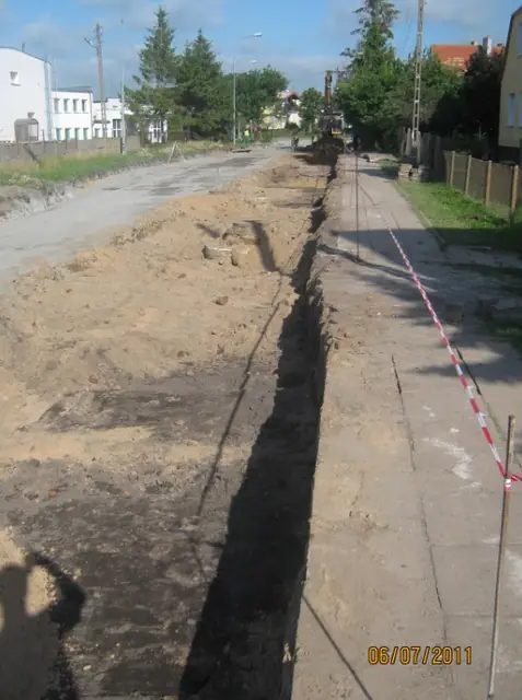 Długi prosty wykop instalacyjny wzdłuż pobocza drogi oznaczony czerwono-białą taśmą