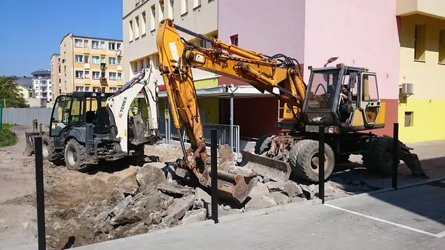 Dwie koparki rozbierają starą nawierzchnię drogi wewnętrznej przy bloku mieszkalnym