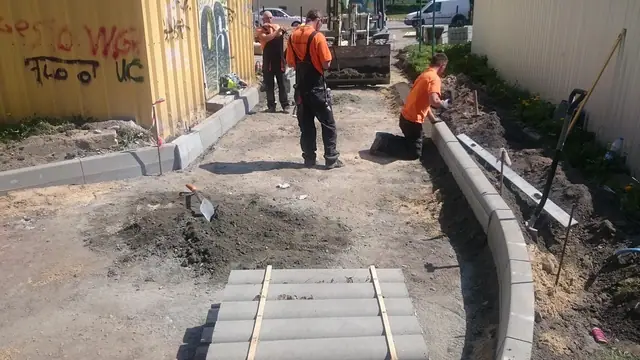 Ekipa układa krawężniki i podbudowę drogi przy żółtej elewacji budynku