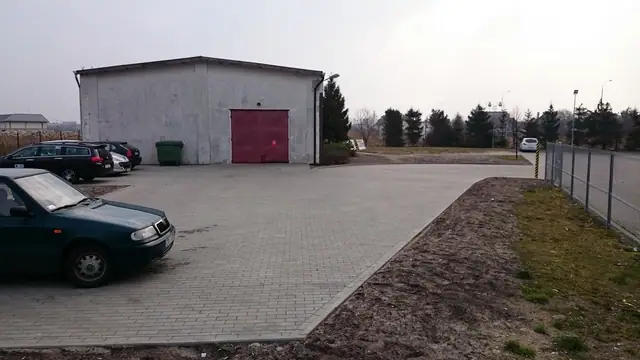Parking z kostki brukowej przy szarym budynku gospodarczym z czerwoną bramą garażową