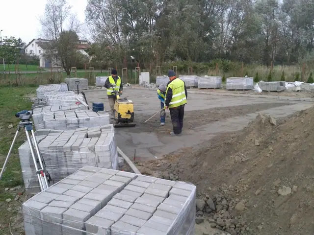 Robotnicy zagęszczają podsypkę obok spiętrzonych palet kostki brukowej na placu