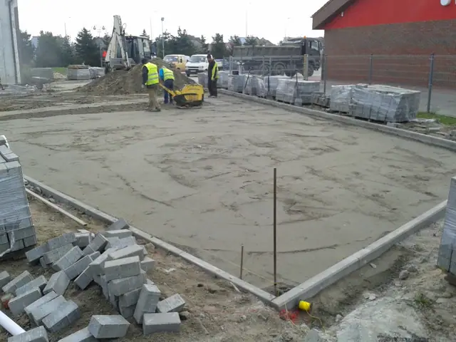 Robotnicy prowadzą zagęszczarkę na piaskowej podsypce obok stosów kostki brukowej i krawężników