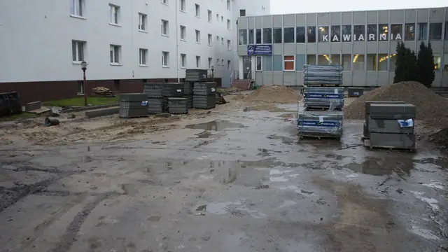 Błotnisty plac między blokami z paletami elementów betonowych i kontenerami na odpady